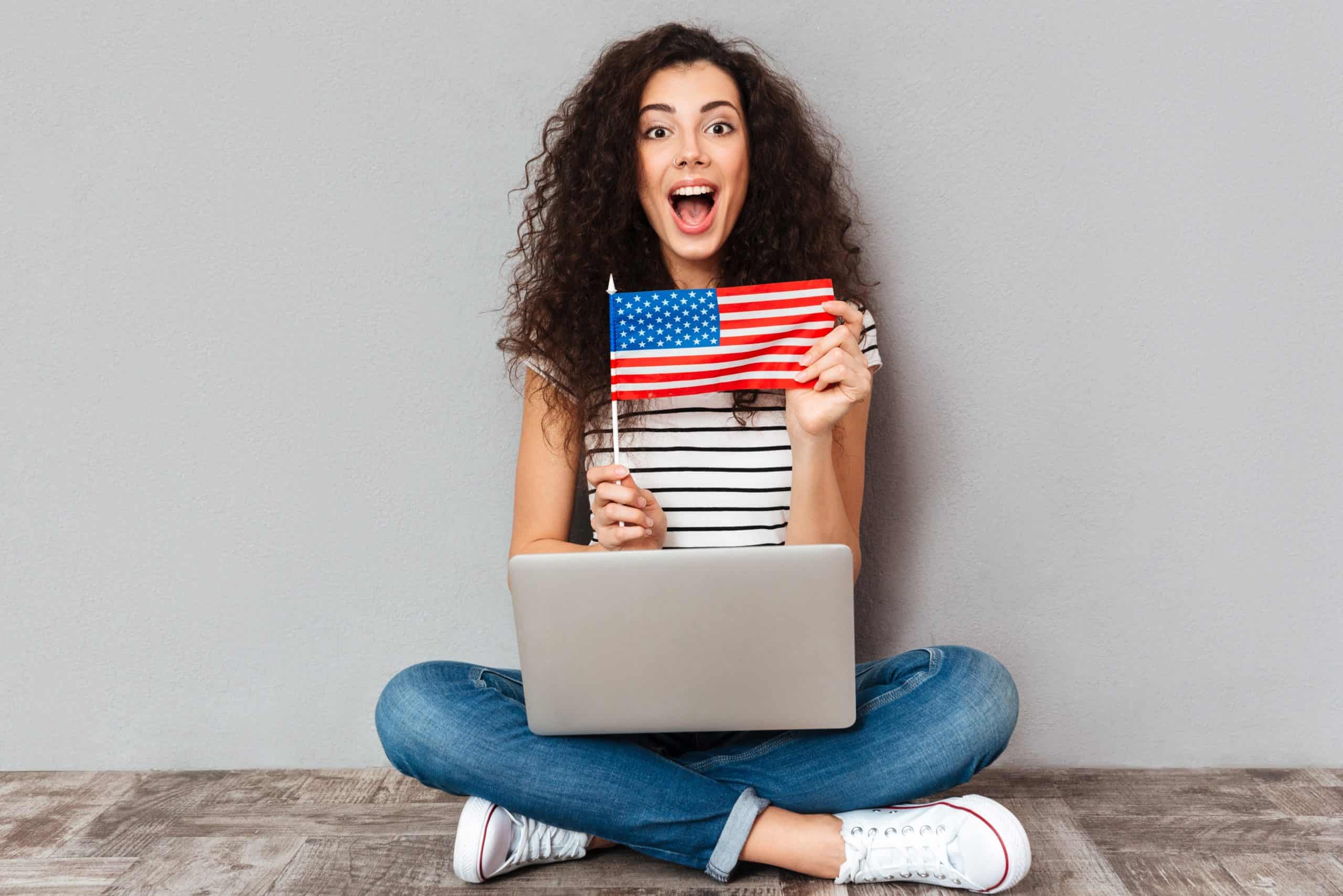 Gorgeous female with beautiful smile sitting in lotus pose with silver computer on legs, demonstrating american flag on camera over grey wall